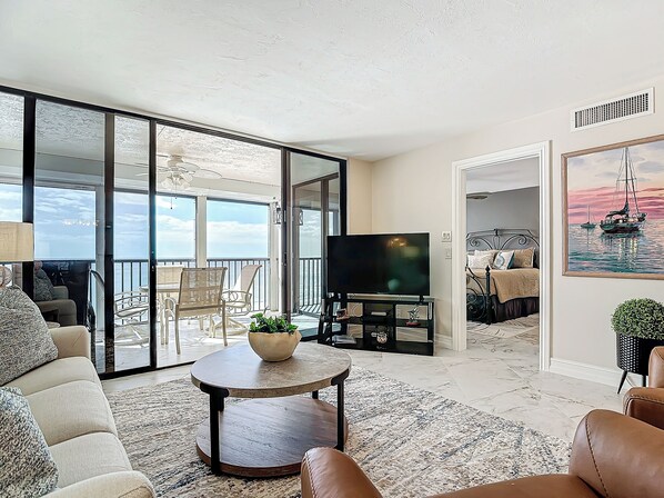 A spacious living area with a large TV, comfortable seating, a balcony with a view, and a painting on the wall.