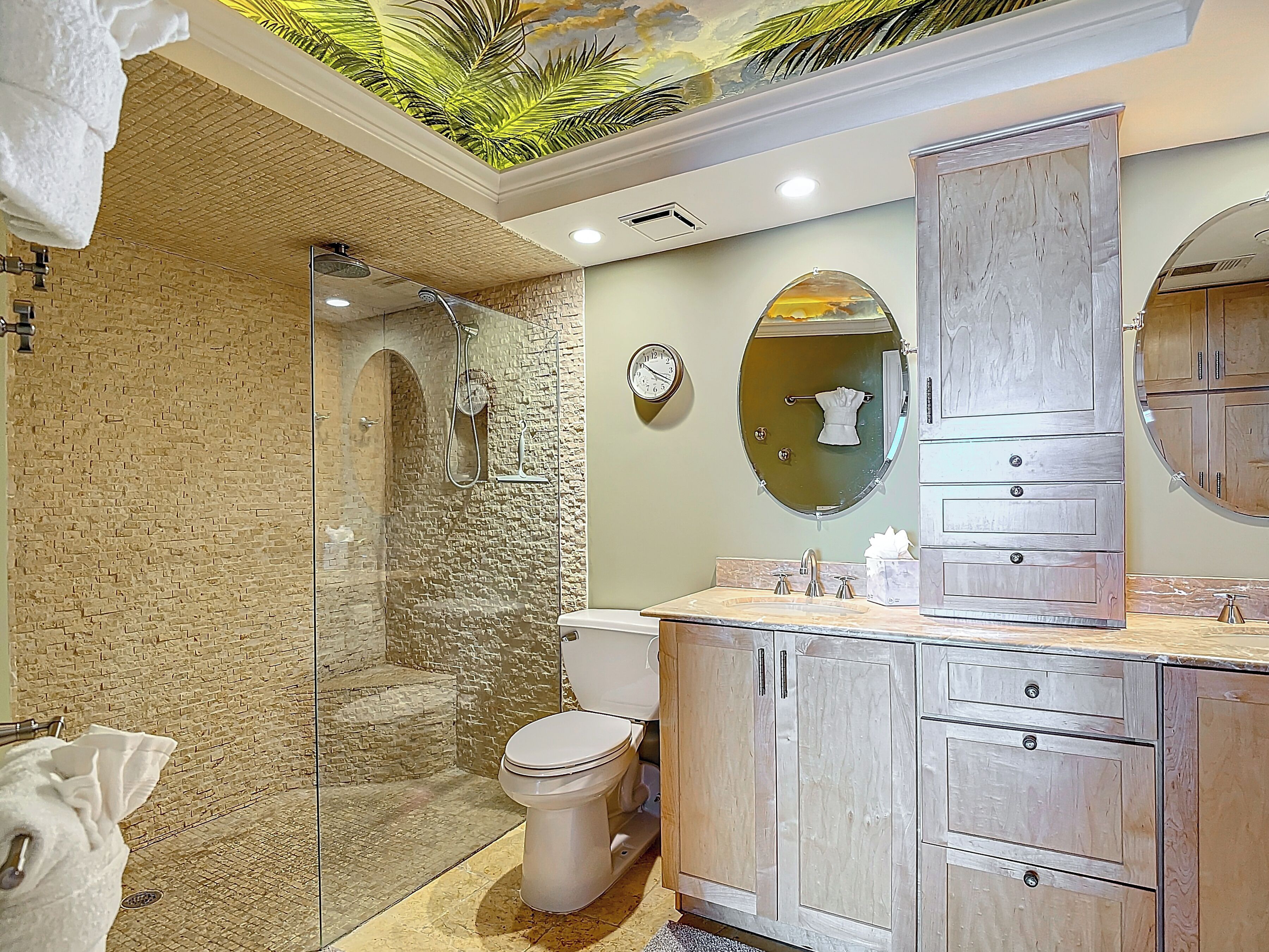 A bathroom with a stone shower enclosure, a toilet, a sink with a mirror, and wooden cabinets.