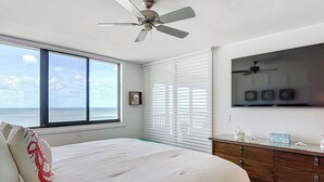 Un dormitorio con una cama grande, un ventilador de techo, vista al océano y un aparador de madera con objetos decorativos.