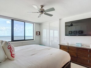 A bedroom with a large bed, a ceiling fan, a view of the ocean, and a wooden dresser with decorative items.