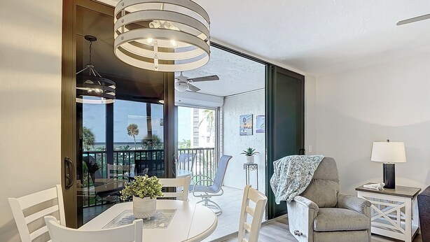 Un moderno comedor con una mesa redonda y sillas blancas, un sillón gris, una mesita auxiliar con una lámpara, y vistas al exterior a través de un ventanal.