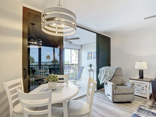 A modern dining area with a round table and white chairs, a grey armchair, a small side table with a lamp, and a view of the outdoors through a glass door.