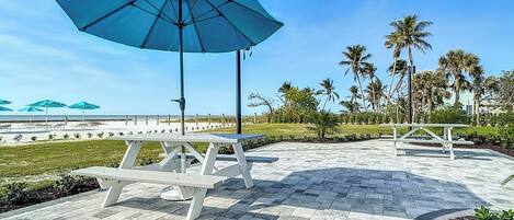 A patio area with picnic tables and umbrellas, surrounded by palm trees and a view of the beach.