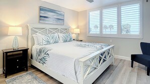 Un dormitorio con cama, ventilador de techo, mesita de noche, lámpara, ventana con persianas y una silla.