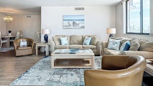Ein Wohnzimmer mit einem beigen Sofa, einem Couchtisch und einem gemusterten Teppich. An der Wand hängt ein Gemälde und das Fenster bietet eine Aussicht.