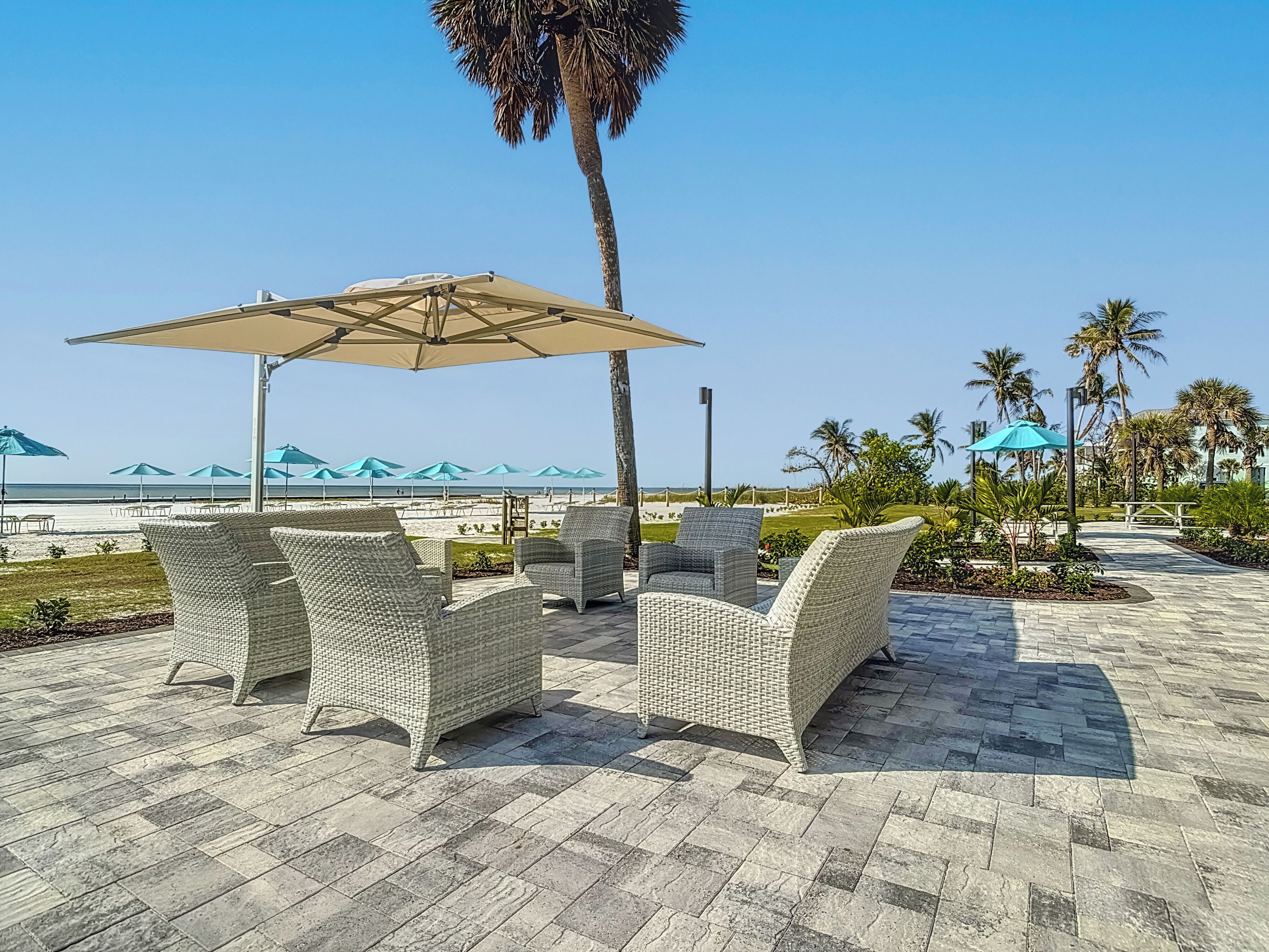 Une terrasse dotée de meubles en osier et de parasols, offrant une vue sur une plage bordée de palmiers et un ciel dégagé.