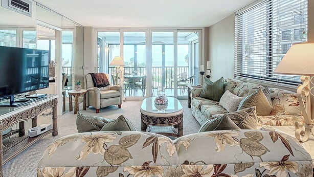 A spacious living room with a large flat-screen TV, comfortable seating, a balcony with outdoor furniture, and large windows.