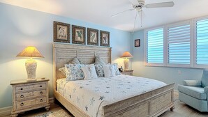 Un dormitorio con cama de madera, dos mesitas de noche con lámparas, un ventilador de techo y una ventana con persianas.