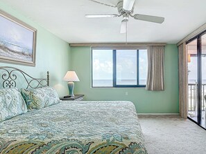 A bedroom with a bed, a ceiling fan, a nightstand with a lamp, a window with curtains, and a view of the sea.