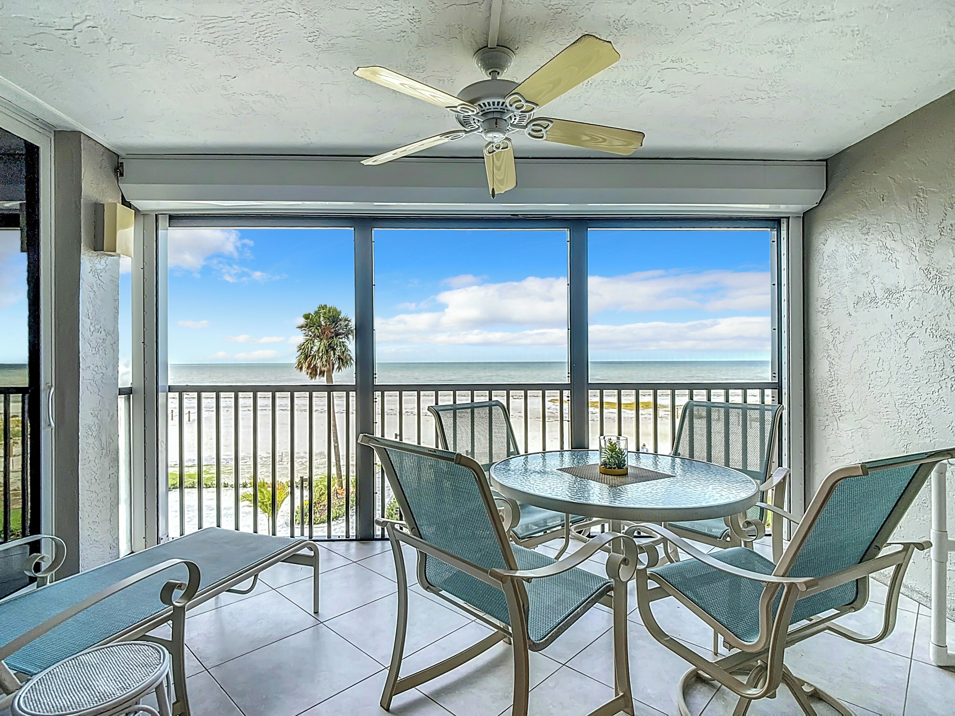 A balcony with a glass table and chairs, a ceiling fan, and a view of the beach.
