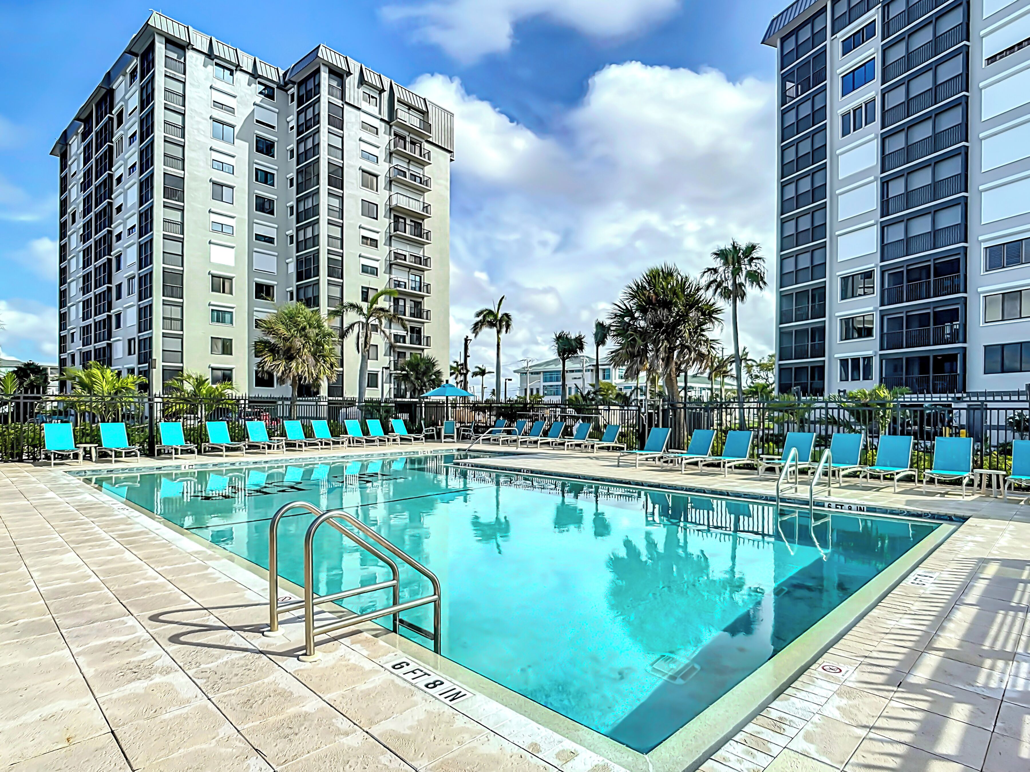 A swimming pool with blue lounge chairs and a metal ladder, surrounded by tall buildings and palm trees.
