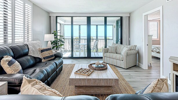 A modern living room with a black leather sofa, striped armchair, and a wooden coffee table. A large sliding glass door leads to an outdoor balcony with a view of the ocean.
