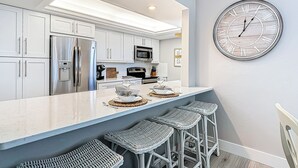 A modern kitchen with white cabinetry, stainless steel appliances, a large island with bar stools, and a wall-mounted clock.