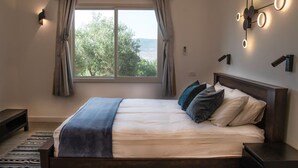 A bedroom with a large bed, a view of trees through the window, and a wall-mounted light fixture. - Charming 2-bedroom apartment with WiFi, AC in quaint Mikhmanim (Mikhmanim)