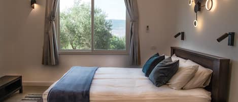 A bedroom with a large bed, a view of trees through the window, and a wall-mounted light fixture.