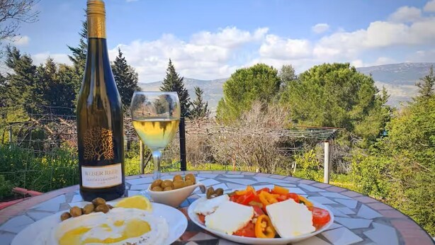 A table with a bottle of wine, a glass of white wine, and plates of food including eggs, olives, cheese, and vegetables. - Charming 2-bedroom apartment with WiFi, AC in quaint Mikhmanim (Mikhmanim)