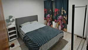 A bedroom with a bed, a nightstand, a standing fan, and a window with curtains.