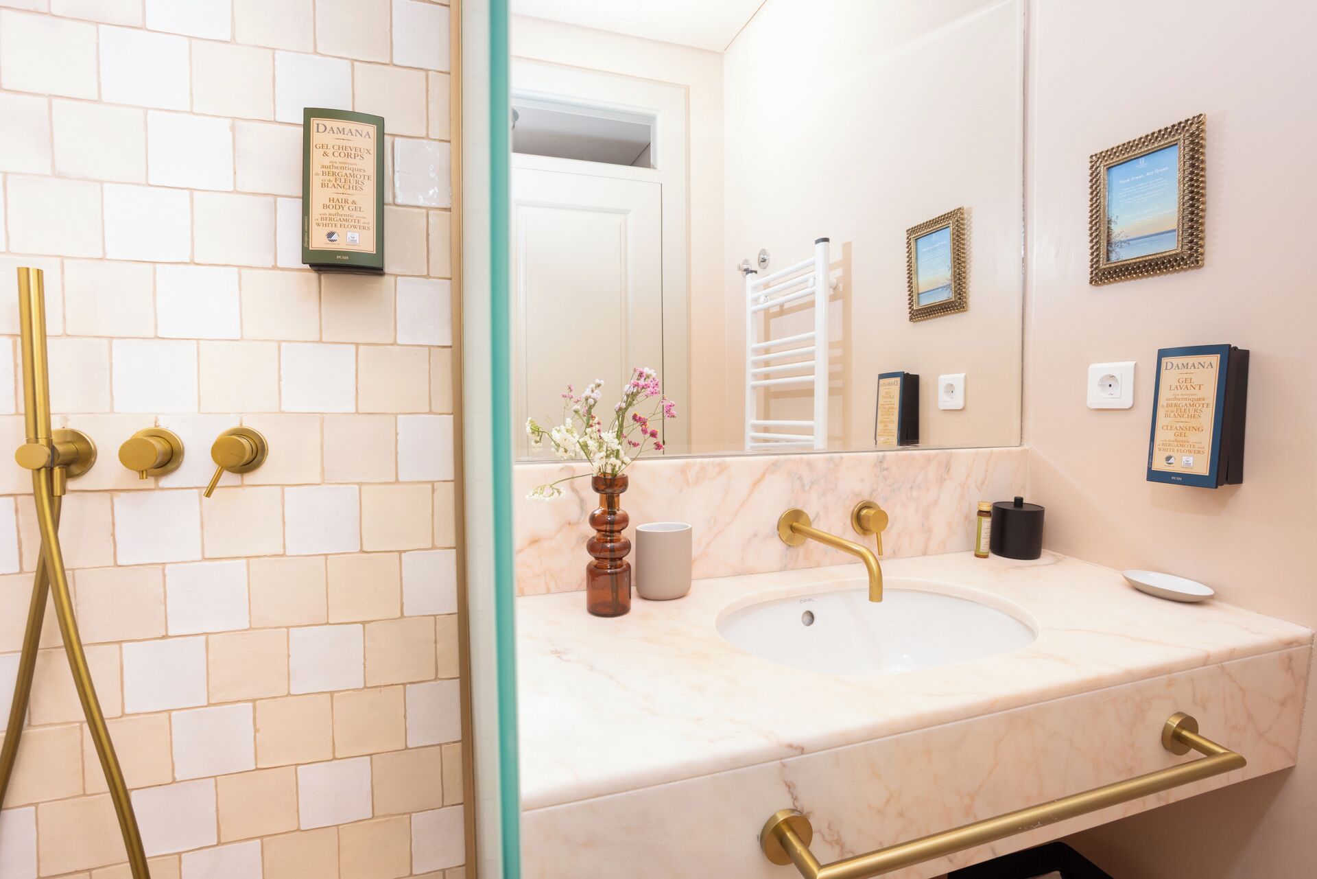 A bathroom with a white tiled shower, a marble sink, and gold fixtures. There are framed pictures on the wall and a vase with flowers on the sink counter.