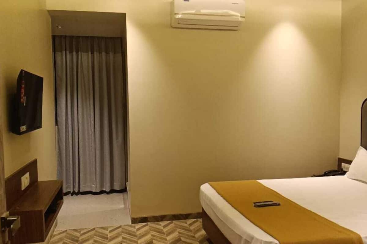 A hotel room with a bed, a bedside table, a TV mounted on the wall, and an air conditioning unit.