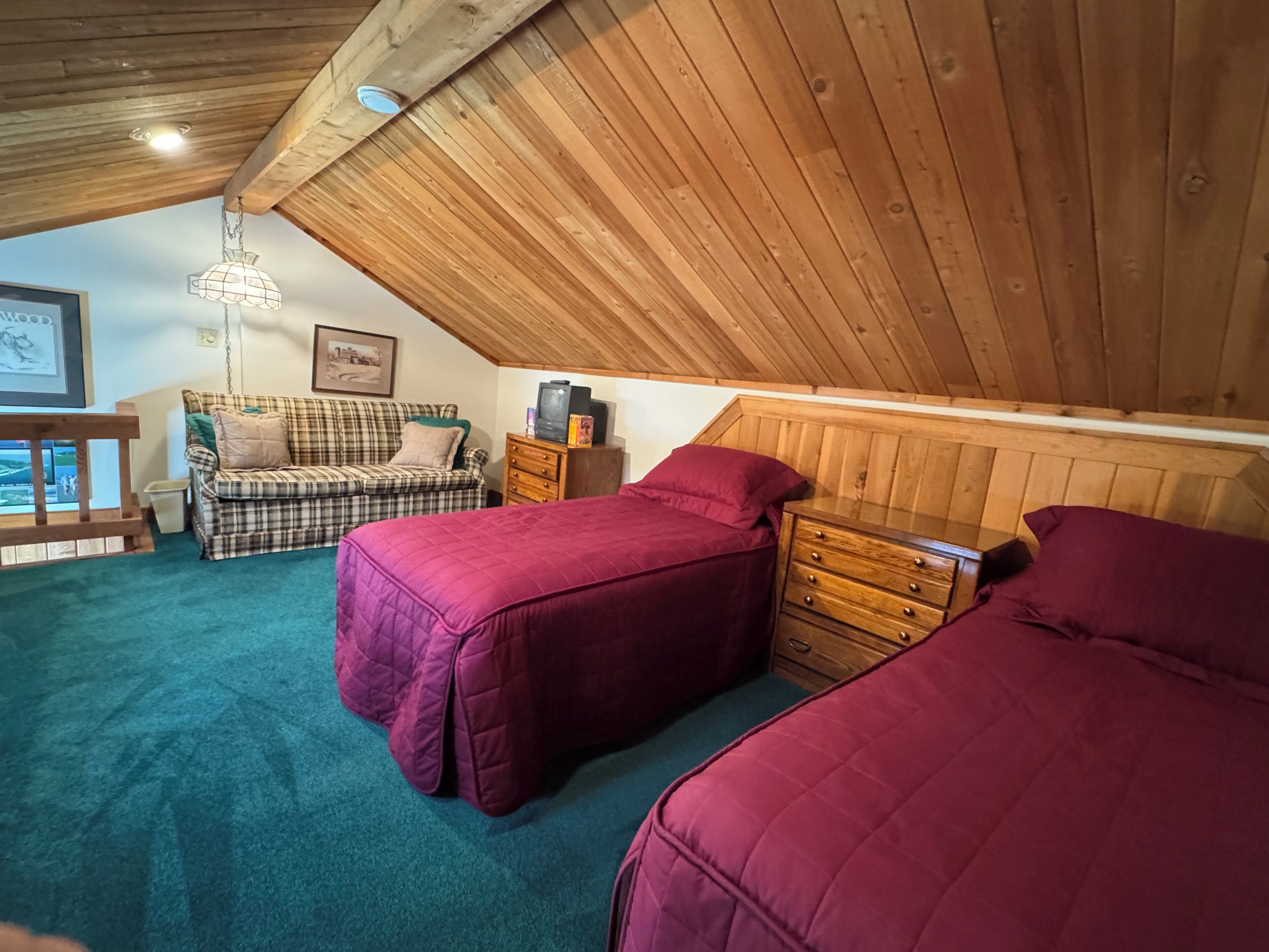 Une chambre confortable située sous les toits, avec un plafond en bois, un canapé à carreaux, deux lits avec une literie rouge et une commode.