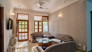 Ein geräumiges Wohnzimmer mit hoher Decke, Deckenventilator und einem großen Fenster mit Blick auf Grünflächen. Zwei Sofas und ein Glastisch sind vorhanden.