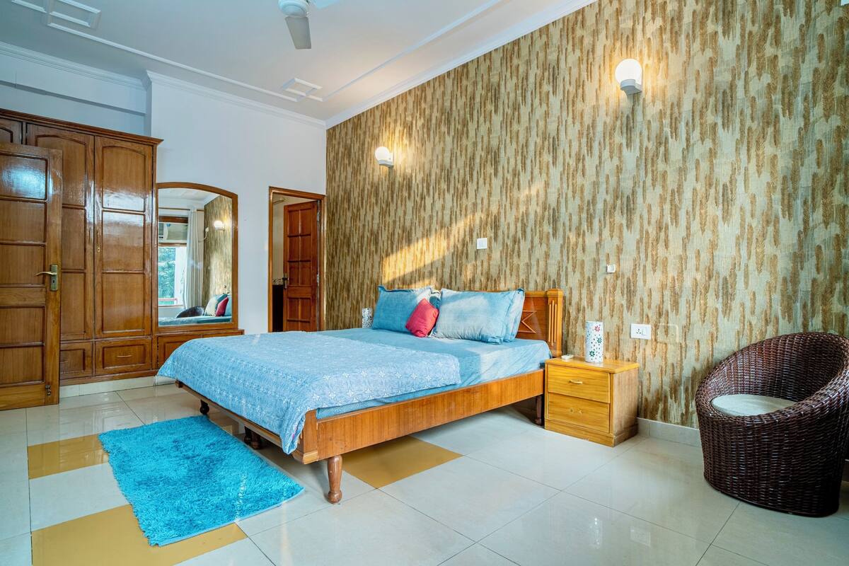Ein Schlafzimmer mit einem hölzernen Bett, einem blauen Teppich, einem Korbstuhl und einer Wand mit Muster.