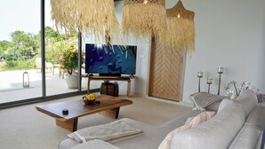 A modern living room with a large flat-screen TV, a wooden coffee table, a grey sofa, and a large window offering a view of greenery. - New single storey villa with sea view, all comforts, close to research area. (Bophut)