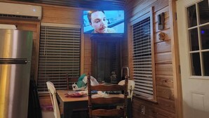 A wooden cabin interior with a dining table, chairs, a television mounted on the wall, and a refrigerator. - Charming 1-bedroom house with WiFi, AC on the Bayou in beautiful Bay St. Louis (Bay St. Louis)