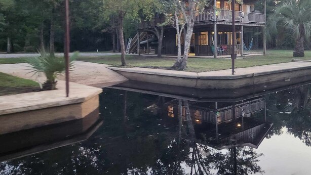 A two-story wooden building with a balcony, surrounded by trees and a calm water body. - Charming 1-bedroom house with WiFi, AC on the Bayou in beautiful Bay St. Louis (Bay St. Louis)