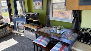 Un espacio habitable compacto con una televisión, una mesita con camino de mesa decorativo, una ventana con cortinas y una repisa con una figurita de pájaro.