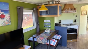 Una cocina compacta con estufa, una mesa de comedor con sillas, un televisor y una ventana con cortinas.