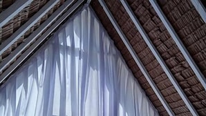 Una habitación triangular con techo inclinado, cortinas blancas y una cama con almohadas.