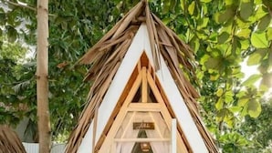 Estructura en forma de tienda de campaña triangular con una cama en su interior, rodeada de follaje natural.
