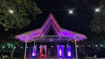 Ein Pavillon mit dreieckigem Dach und einer Bühne mit gemustertem Hintergrund, umgeben von Bäumen und Lichterketten.