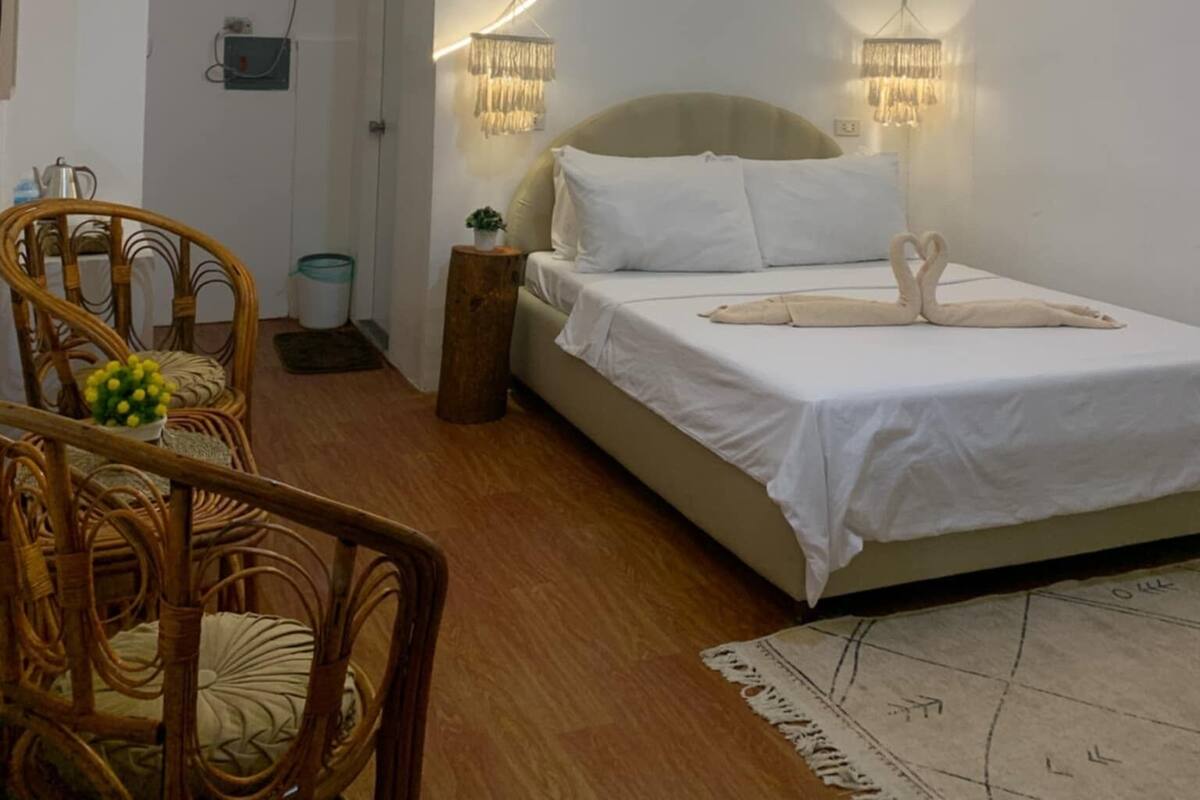 A modern bedroom with a bed, two bedside lamps, a ceiling light, a wicker chair, and a small table with a plant.