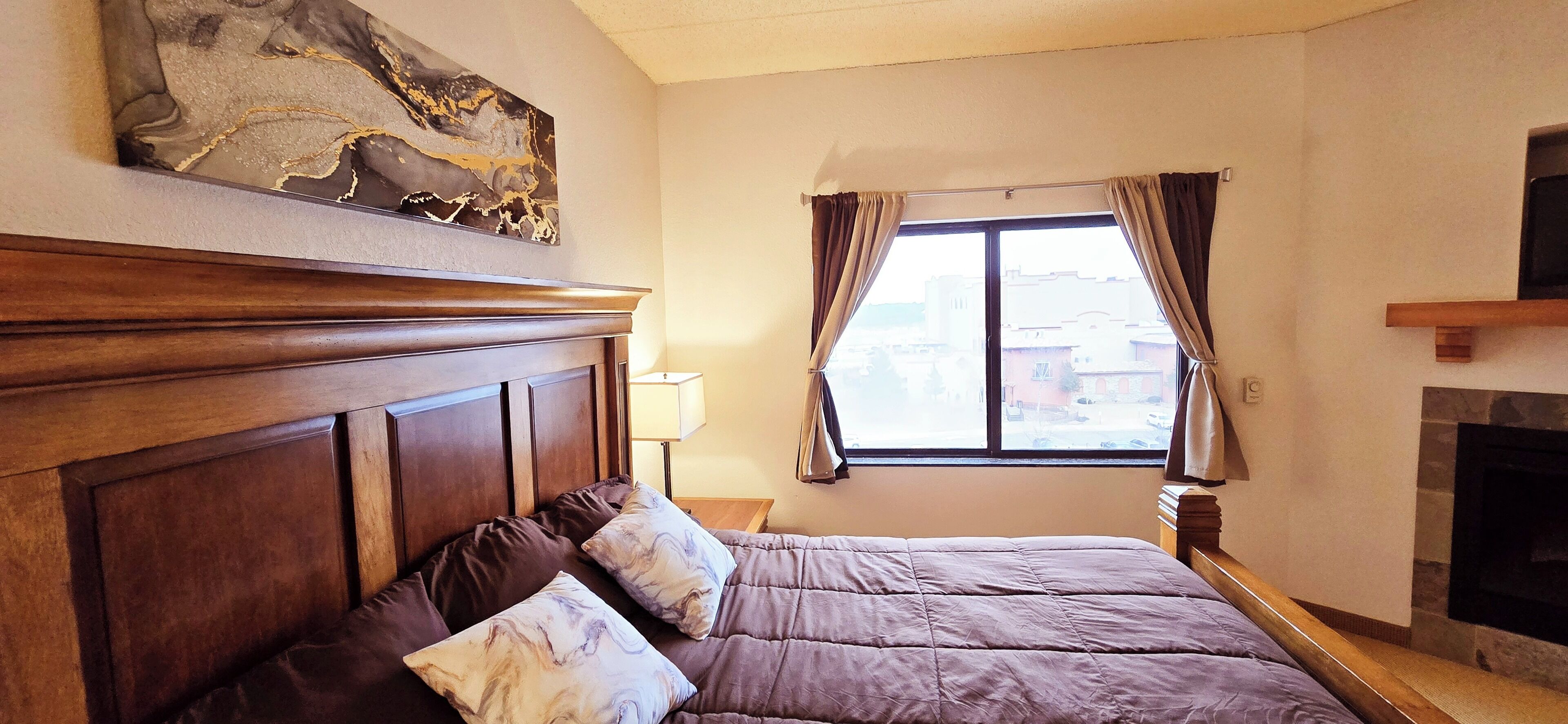 A bedroom with a large bed, a wooden headboard, a fireplace, a window with curtains, and a painting on the wall.