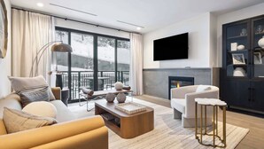 A modern living room with a sofa, coffee table, and armchairs, a fireplace, and a large window with a view of snow-covered trees.
