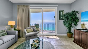 A modern living room with a glass coffee table, a sofa, a chair, a lamp, a large potted plant, a painting, and a view of the ocean through a sliding glass door.