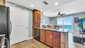 A modern kitchen with wooden cabinets, a black refrigerator, and a dishwasher. There is a dining area with a table and chairs, and a view of the ocean through large windows.