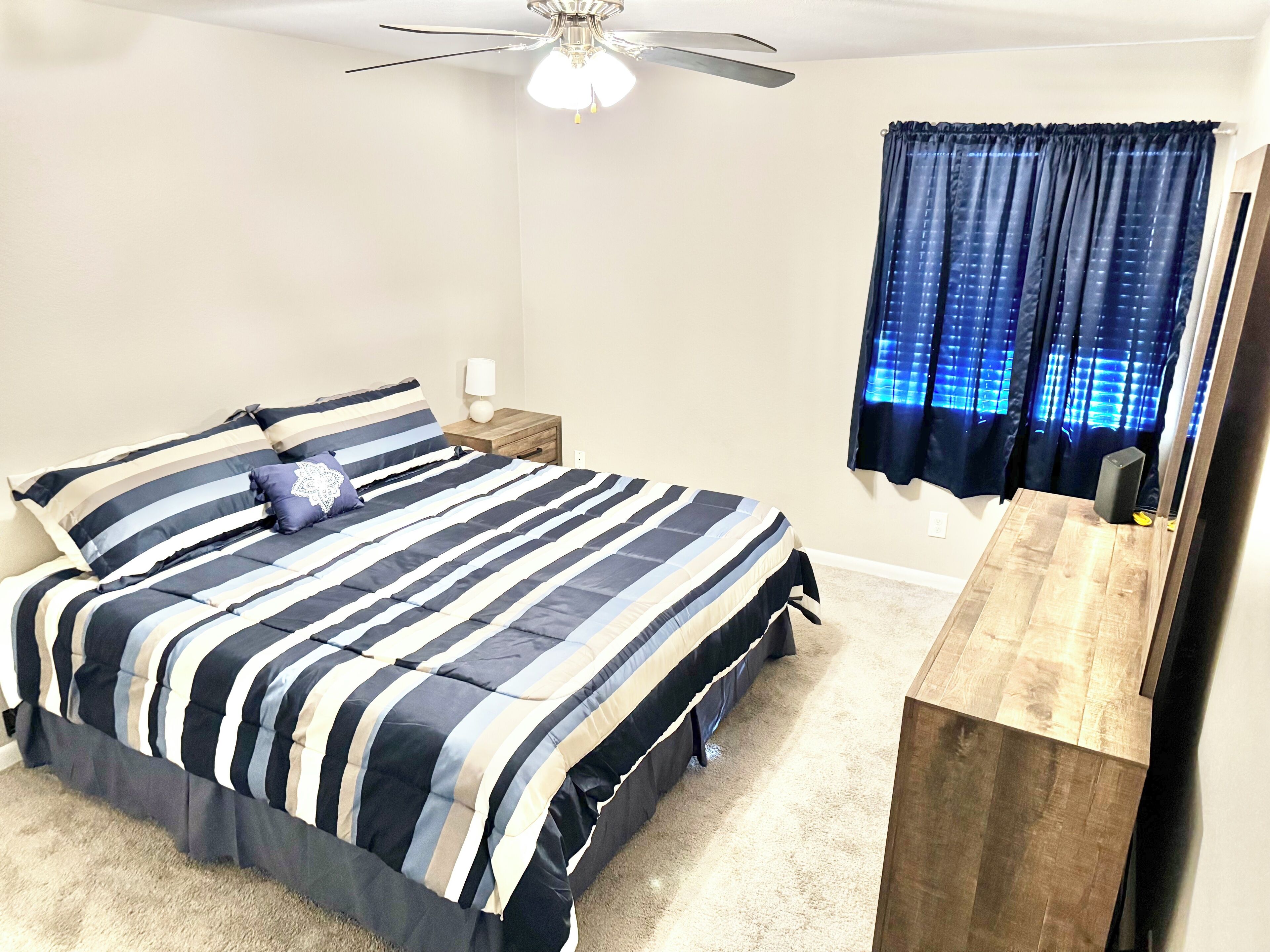A bedroom with a bed, a nightstand, a ceiling fan, a window with curtains, and a wooden dresser.