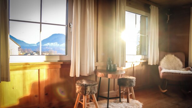 A cozy room with wooden walls, a wooden floor, a window with a mountain view, a small table with a vase and cup, and a fluffy chair. - Quaint vacation home in the mountains - pure relaxation and nature! (Lingenau)