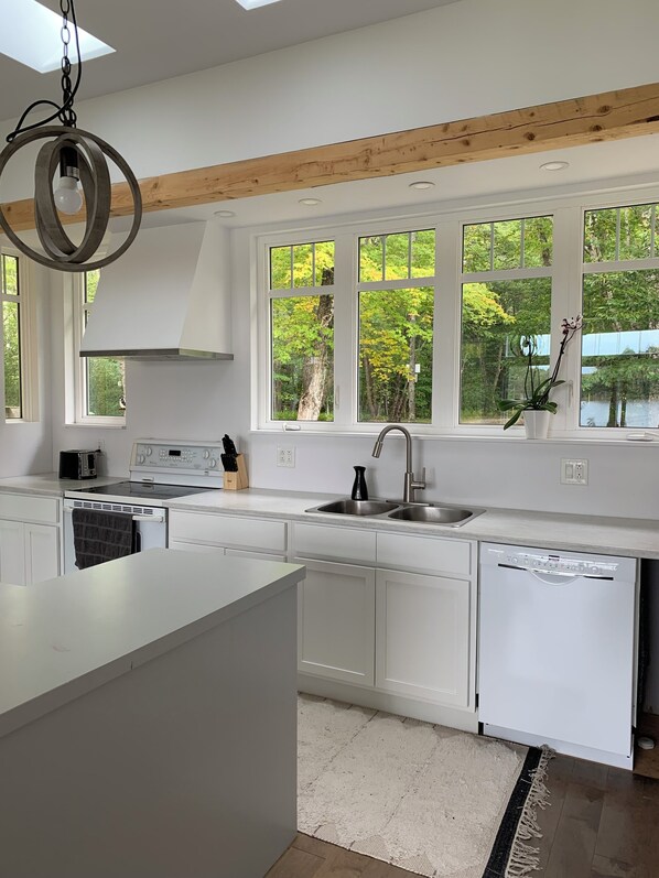 A modern kitchen with white cabinetry, a built-in dishwasher, a stainless steel sink, and a large window with a view of trees. - Private home on 8 acres of wooded and flower-filled grounds at Wentworth Lake (Moodys Corner)