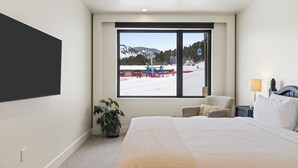 A hotel room with a large bed, a potted plant, a chair, a nightstand, and a window with a view of a ski lift and mountains.