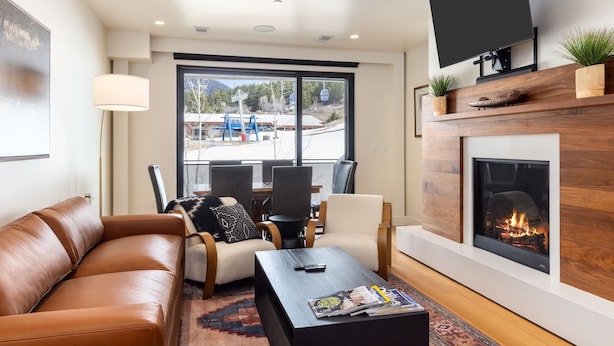A modern living room with a leather sofa, a coffee table, and a fireplace. There is a TV mounted above the fireplace and a large window providing a view of the outdoors.
