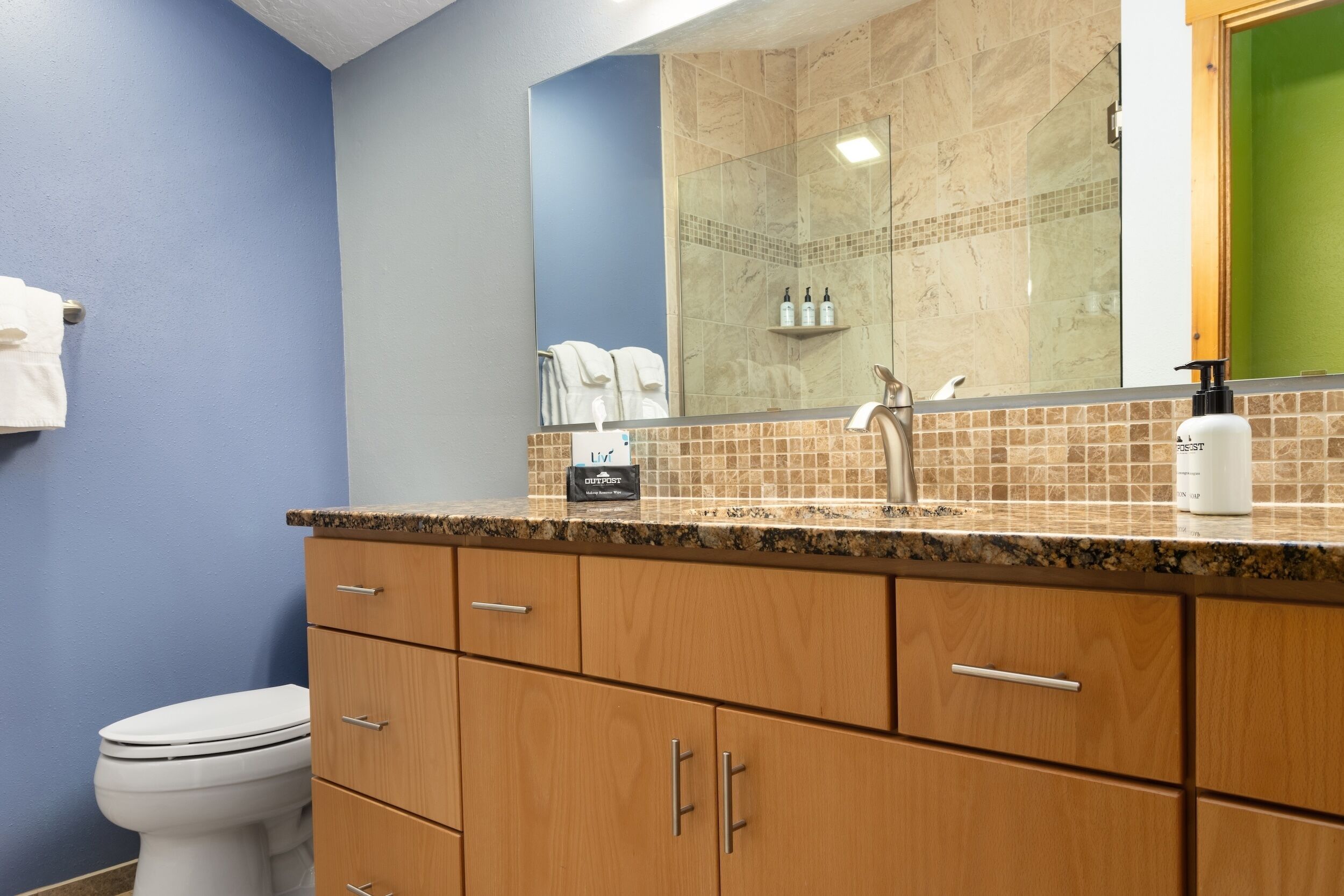 A bathroom with a white toilet, wooden cabinets, a granite countertop, a mirror, and a shower area.