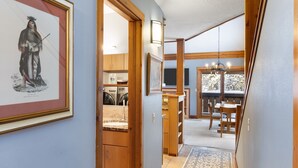 Un couloir avec des meubles de rangement en bois, un tableau encadré, un lustre et un coin repas comprenant une table et des chaises.