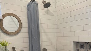 A bathroom with a white tiled shower, a wooden vanity with a sink, a mirror, and a decorative tile backsplash.