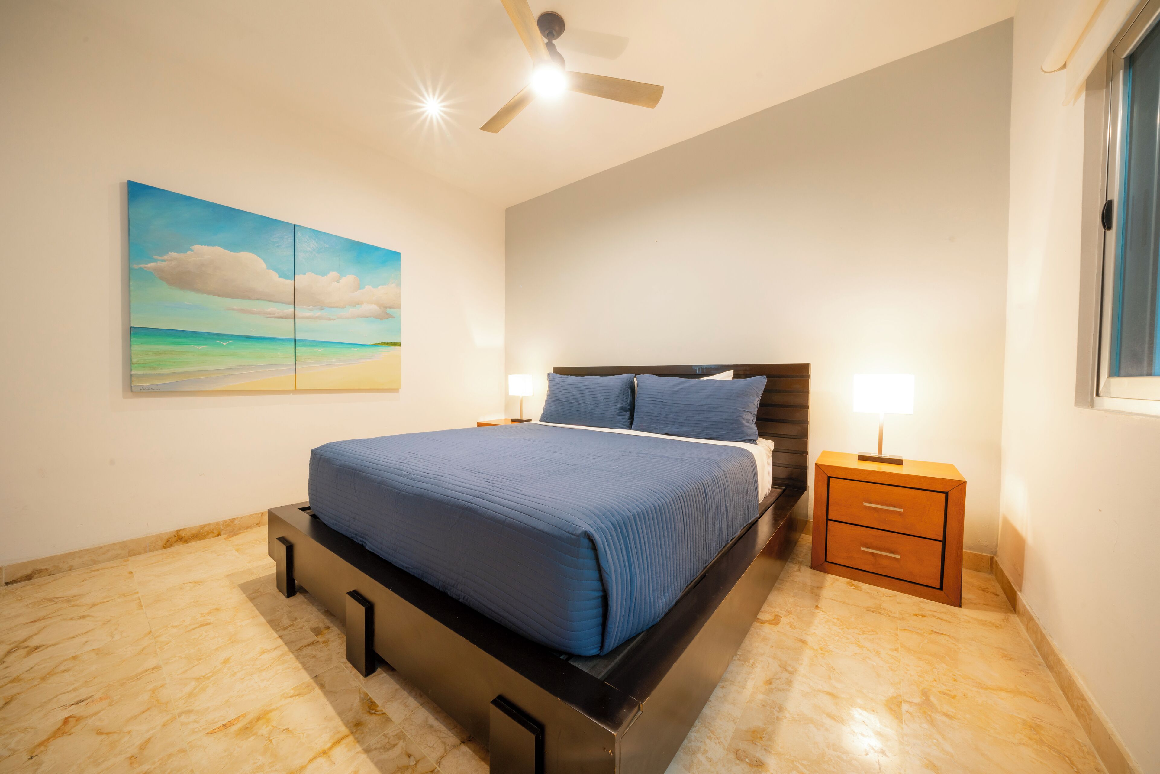 Une chambre à coucher avec un lit, des tables de chevet, un ventilateur de plafond et un tableau représentant une scène de plage.