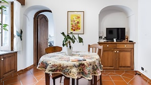 Un comedor con mantel floral, sillas de madera, una planta en maceta y un televisor sobre un mueble de madera.