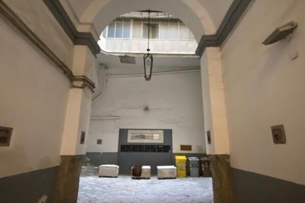 A corridor with a high ceiling, a hanging light fixture, and a window with bars. There are several white objects on the floor, possibly furniture or equipment.